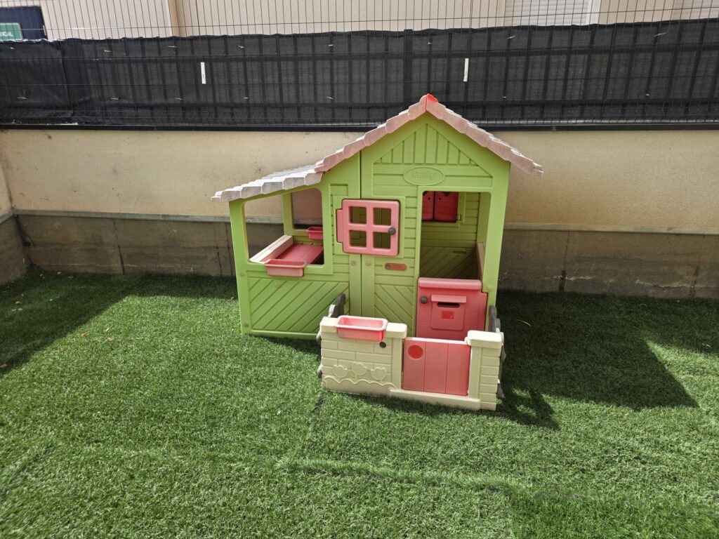 Espace extérieur de la micro crèche avec de la pelouse synthétique et une maisonnette pour enfants verte et rose